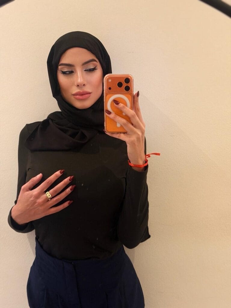 Woman in a black hijab and top taking a mirror selfie with an orange phone, showing red nails and a ring.