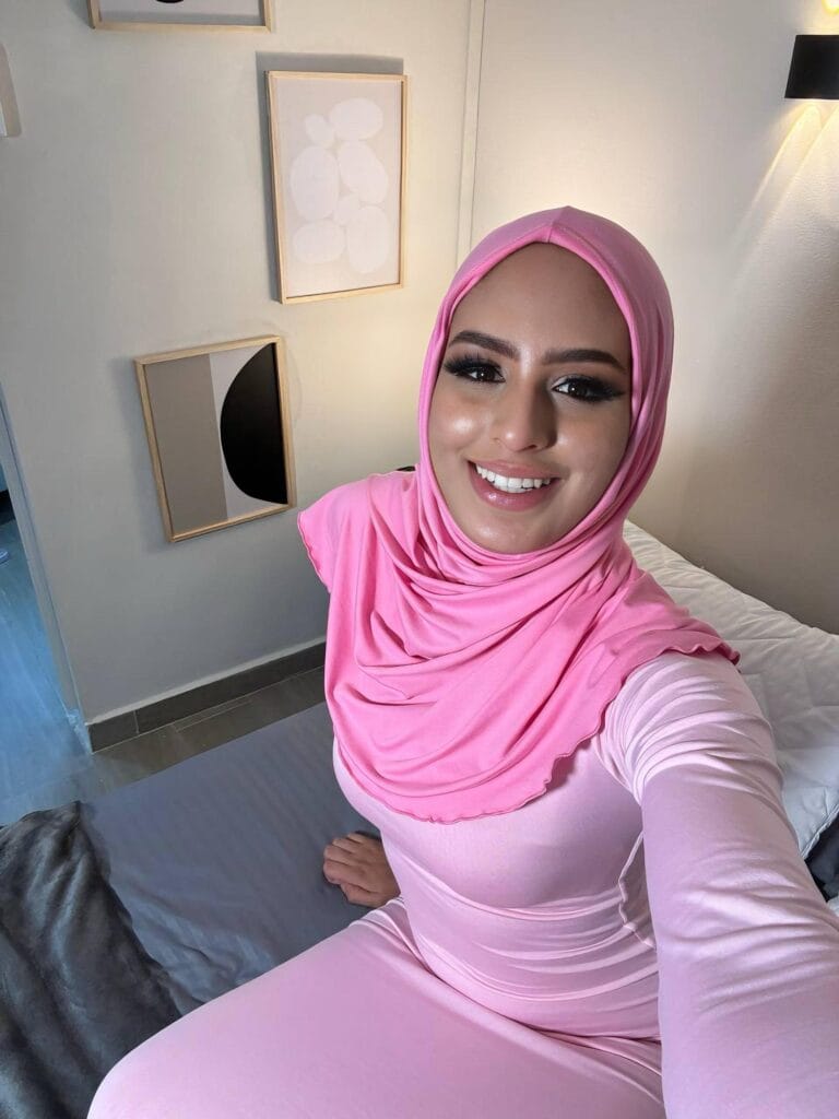 Smiling woman in a pink hijab and matching top taking a selfie in a minimalist room with framed art on the wall.