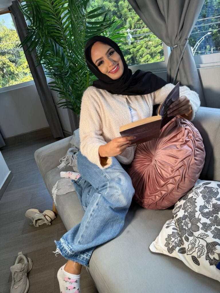 Woman wearing a black hijab sits on a gray sofa, smiling while reading a book indoors near a green plant.