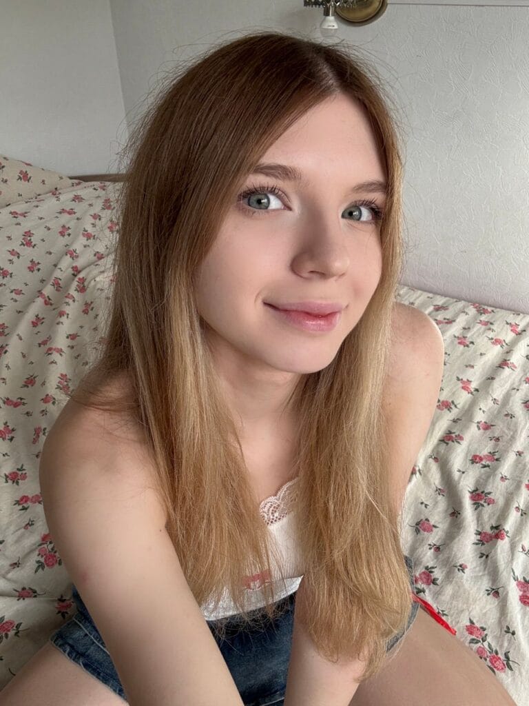 Young woman with long light brown hair and blue-green eyes smiles at the camera, seated on a floral bedspread.