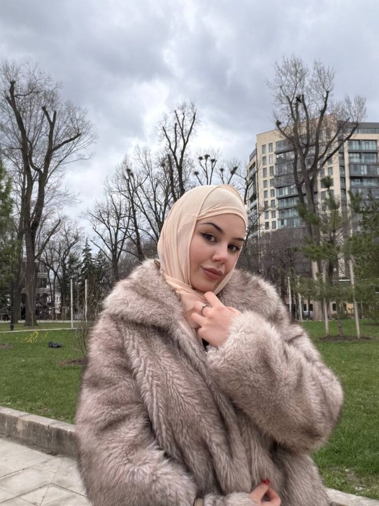 Woman in a beige hijab and fluffy fur coat posing in a park with bare trees and a modern building in the background on a cloudy day.
