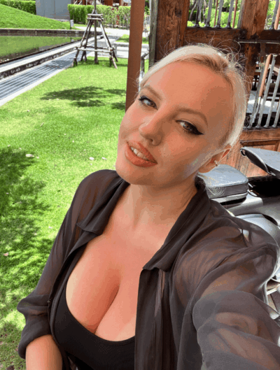Close-up portrait of a blonde woman wearing a black top and sheer black cover-up, posing outdoors in a sunny green garden with wooden structures in the background.