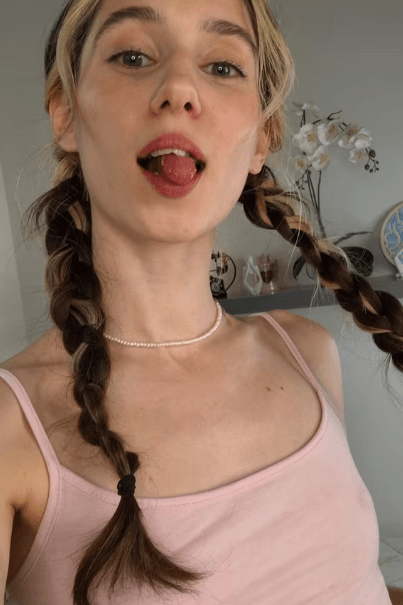 Young woman with two braids wearing a pink tank top, sticking out her tongue playfully in a bright room with orchids in the background